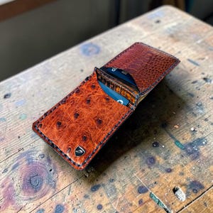 May include: A cognac-colored leather wallet with a textured ostrich-skin pattern, featuring dark blue contrast stitching along the edges, a small silver-toned metal emblem on the front, and a single card slot containing a blue credit card.