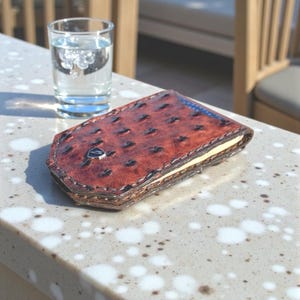 May include: A brown leather wallet with a textured ostrich pattern and a silver emblem. The wallet is open, revealing a cream-colored interior. A glass of water sits next to the wallet.