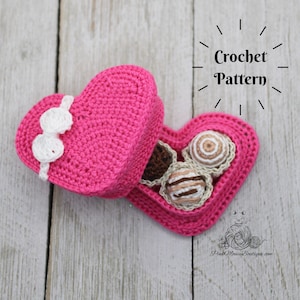 Könnte beinhalten: Eine rosa gehäkelte herzförmige Schachtel mit einer weißen gehäkelten Schleife. Die Schachtel ist geöffnet und enthält drei gehäkelte Schokoladenbonbons.