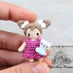 May include: A small crocheted doll with brown hair, white bows, and a pink dress. The doll is holding a small crocheted animal in its arms.