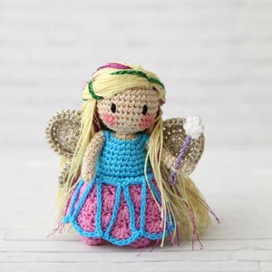 Puede incluir: Una mu&ntilde;eca de hada de crochet con cabello rubio, un vestido azul, una falda rosa y una varita blanca. La mu&ntilde;eca lleva una corona de flores verde y morada.