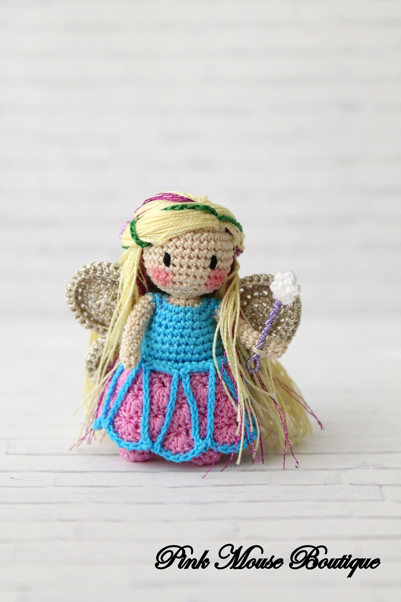 Puede incluir: Una mu&ntilde;eca de hada de crochet con cabello rubio, un vestido azul, una falda rosa y una varita blanca. La mu&ntilde;eca lleva una corona de flores verde y morada.