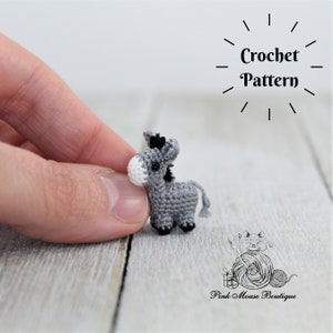 May include: Crochet pattern for a miniature gray donkey with black hooves and a white muzzle. The donkey is being held in a hand.