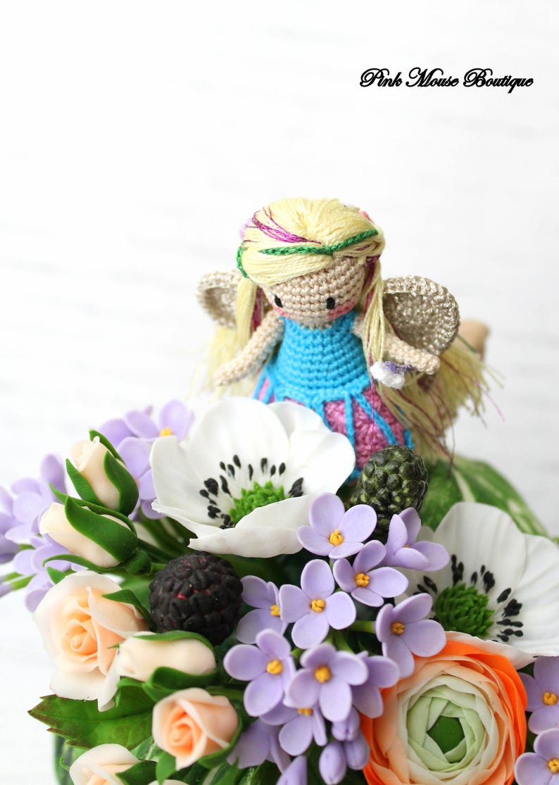 Puede incluir: Una mu&ntilde;eca de hada de crochet con cabello rubio y alas azules se sienta encima de un ramo de flores blancas, rosas y moradas. Las flores est&aacute;n hechas de arcilla polim&eacute;rica e incluyen rosas, an&eacute;monas y lilas.