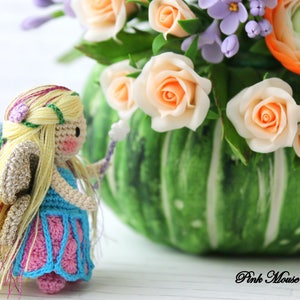 Puede incluir: Una mu&ntilde;eca de hada de crochet con cabello largo y rubio, vestida con un vestido azul y rosa, sosteniendo una varita. La mu&ntilde;eca est&aacute; de pie frente a un jarr&oacute;n verde con un ramo de flores, incluyendo rosas y ran&uacute;nculos.