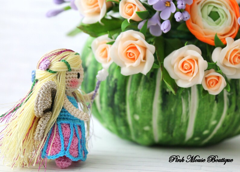 Puede incluir: Una mu&ntilde;eca de hada de crochet con cabello largo y rubio, vestida con un vestido azul y rosa, sosteniendo una varita. La mu&ntilde;eca est&aacute; de pie frente a un jarr&oacute;n verde con un ramo de flores, incluyendo rosas y ran&uacute;nculos.
