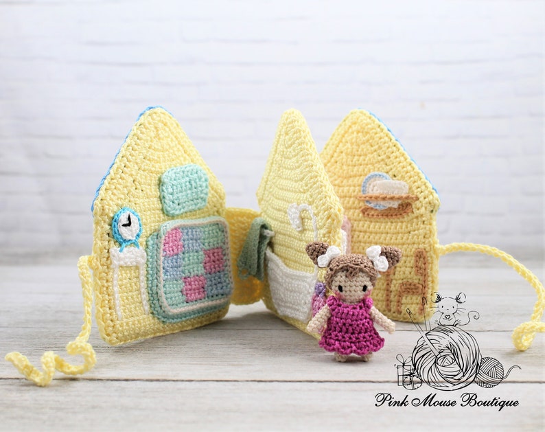May include: A yellow crocheted dollhouse with four rooms, each with a different scene. The dollhouse is open to reveal the interior. A small crocheted doll in a pink dress stands in front of the dollhouse.