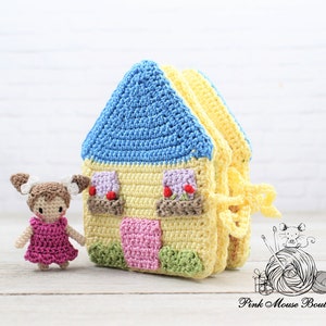 May include: A crocheted house toy with a blue roof, yellow walls, and pink and green details. A small crocheted doll with brown hair and a pink dress stands next to the house.