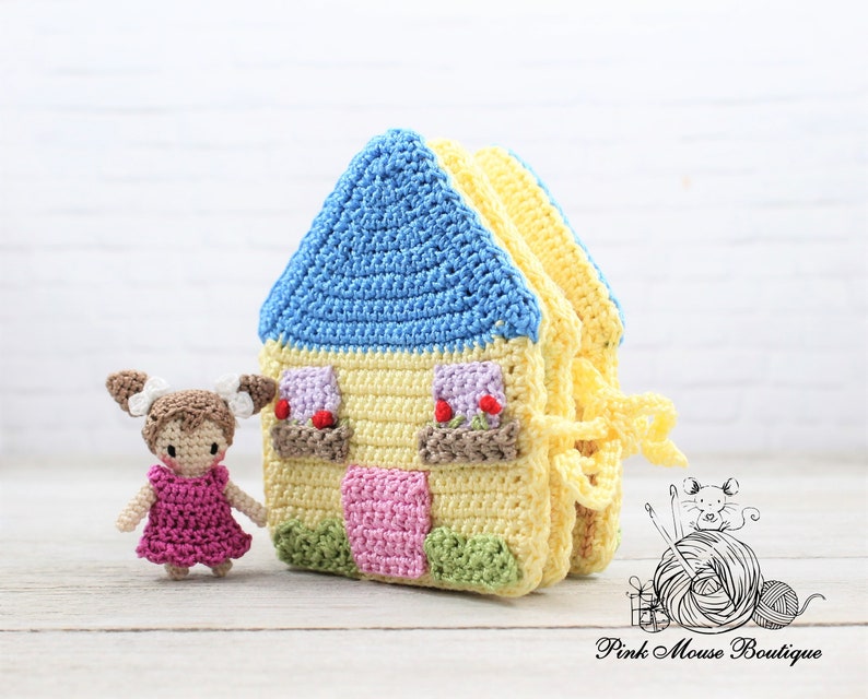 May include: A crocheted house toy with a blue roof, yellow walls, and pink and green details. A small crocheted doll with brown hair and a pink dress stands next to the house.