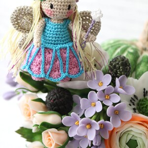 Puede incluir: Una mu&ntilde;eca de hada de crochet con cabello rubio, un vestido azul y alas rosadas, sentada sobre un ramo de flores. Las flores incluyen rosas blancas, lilas moradas y una ran&uacute;nculo de color melocot&oacute;n.