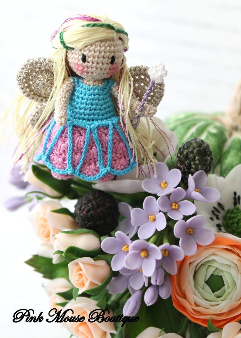 Puede incluir: Una mu&ntilde;eca de hada de crochet con cabello rubio, un vestido azul y alas rosadas, sentada sobre un ramo de flores. Las flores incluyen rosas blancas, lilas moradas y una ran&uacute;nculo de color melocot&oacute;n.