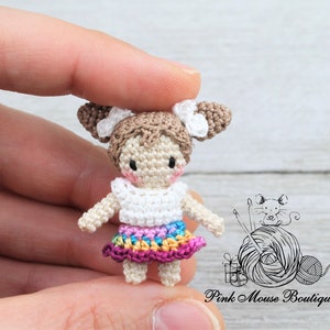 May include: A small crocheted doll with brown hair, white top, and a colorful skirt. The doll is wearing a white crocheted headband with a bow. The doll is being held by a hand.
