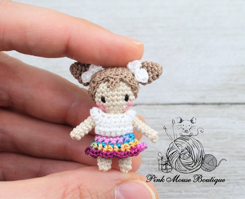 May include: A small crocheted doll with brown hair, white top, and a colorful skirt. The doll is wearing a white crocheted headband with a bow. The doll is being held by a hand.