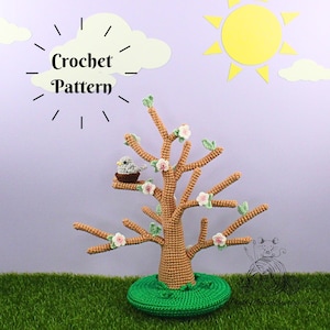 Può includere: Modello a uncinetto per un porta gioielli a forma di albero con un nido di uccello e fiori. Il porta gioielli è marrone e i fiori sono rosa. La base del porta gioielli è verde.