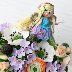 Puede incluir: Una mu&ntilde;eca de hada de crochet con cabello rubio y un vestido azul y rosa, sentada sobre un ramo de flores blancas, rosas y moradas. Las flores est&aacute;n hechas de arcilla polim&eacute;rica e incluyen an&eacute;mones, rosas y ran&uacute;nculos.