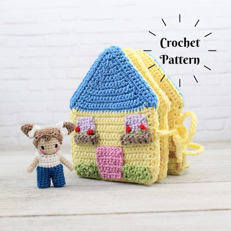 May include: A crocheted house-shaped toy with a blue roof, yellow walls, and a pink door. The house is closed and has a yellow ribbon tied around it. A small crocheted doll with brown hair and a blue dress is standing next to the house. The image includes the text "Crochet Pattern".
