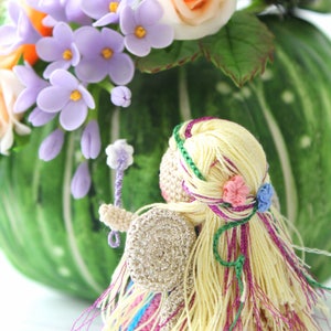 Puede incluir: Una mu&ntilde;eca de hada de crochet con cabello largo rubio, vistiendo un vestido rosa y azul y alas plateadas. La mu&ntilde;eca sostiene una peque&ntilde;a varita y est&aacute; rodeada de flores.