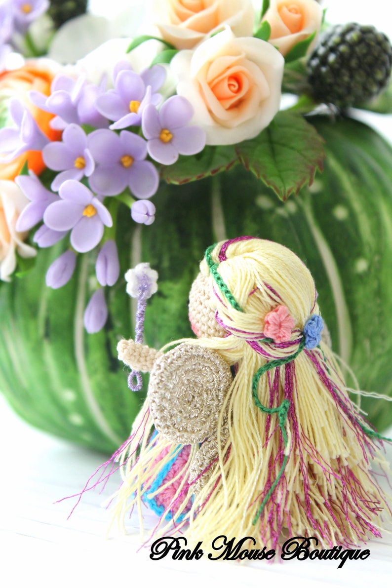 Puede incluir: Una mu&ntilde;eca de hada de crochet con cabello largo rubio, vistiendo un vestido rosa y azul y alas plateadas. La mu&ntilde;eca sostiene una peque&ntilde;a varita y est&aacute; rodeada de flores.