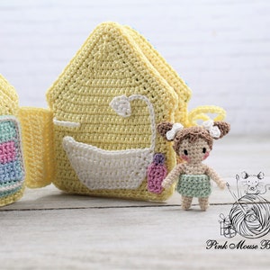 May include: A crocheted play set with a yellow house with a blue and green checkered pattern on the front and a yellow house with a white bathtub and a pink bottle on the front. A small crocheted doll with brown hair and a green dress is standing in front of the houses.