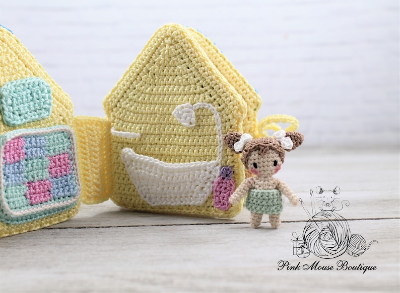May include: A crocheted play set with a yellow house with a blue and green checkered pattern on the front and a yellow house with a white bathtub and a pink bottle on the front. A small crocheted doll with brown hair and a green dress is standing in front of the houses.