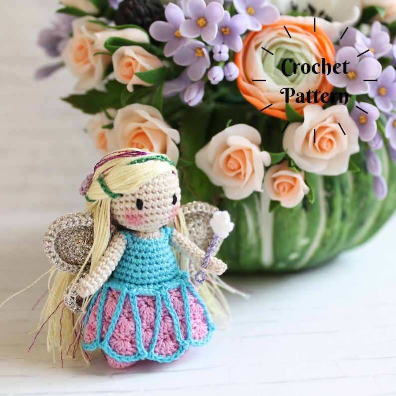 Puede incluir: Una mu&ntilde;eca de hada de crochet con cabello rubio, vestido azul y rosa y alas de purpurina plateada. La mu&ntilde;eca sostiene una varita p&uacute;rpura con una estrella blanca en el extremo. La mu&ntilde;eca est&aacute; sentada sobre una superficie de madera blanca frente a un jarr&oacute;n de flores.