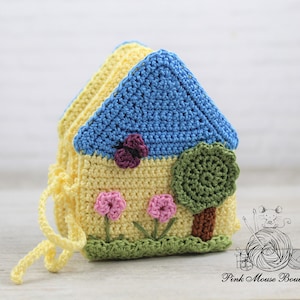 May include: A crocheted house-shaped toy with a blue roof, yellow walls, green grass, pink flowers, and a green tree. The toy has a yellow drawstring.