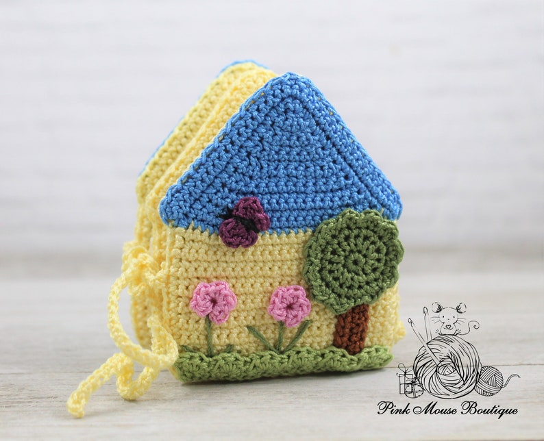 May include: A crocheted house-shaped toy with a blue roof, yellow walls, green grass, pink flowers, and a green tree. The toy has a yellow drawstring.