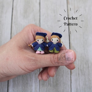 May include: Two crocheted graduation figures, one in a blue gown and green skirt, the other in a blue gown and green trousers, holding hands. The figures are wearing graduation caps and have blonde hair. The text "Crochet Pattern" is above the figures.