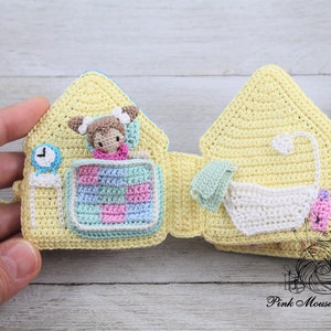 May include: A yellow crocheted house-shaped book with different scenes inside. The first scene shows a doll in bed with a blue and pink quilt. The second scene shows a white bathtub with a green towel and a pink bottle.