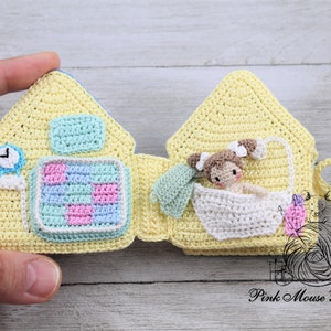 May include: A crocheted yellow house-shaped book with a clock, a checkered pattern, and a doll in a bathtub on the inside pages. The book is open and the doll is visible. The book is made by Pink Mouse Boutique.