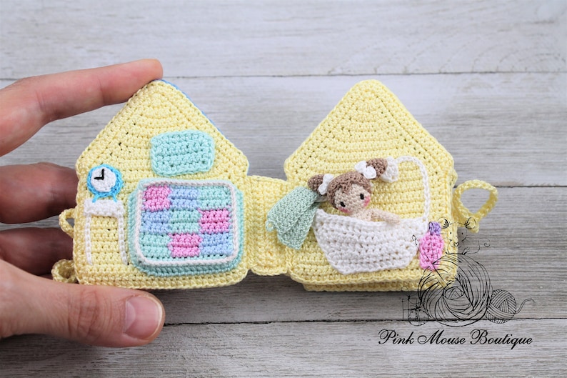 May include: A crocheted yellow house-shaped book with a clock, a checkered pattern, and a doll in a bathtub on the inside pages. The book is open and the doll is visible. The book is made by Pink Mouse Boutique.