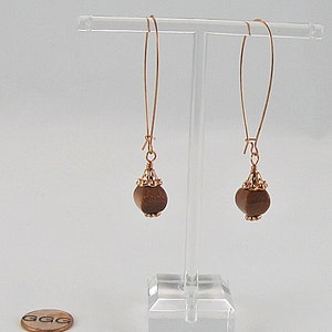 Op de afbeelding: Een paar lange, bruine houten kralen oorbellen met gouden accenten die aan een transparante standaard hangen.