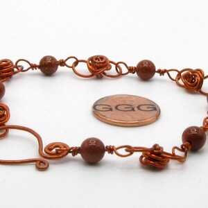 May include: A copper wire bracelet with brown beads and a heart-shaped clasp. The bracelet is about the size of a US penny.