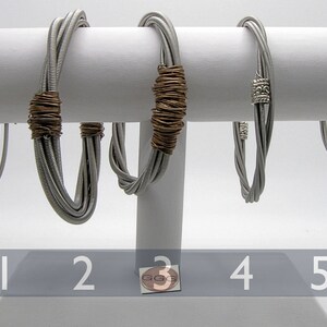 Könnte beinhalten: Fünf silberne Gitarrensaiten-Armbänder mit verschiedenen Verzierungen. Die Armbänder werden auf einem weißen Ständer präsentiert. Die Armbänder sind von 1 bis 5 nummeriert.