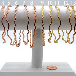May include: Fifteen wavy metal bangles displayed on a white stand. The bangles are in various colors, including gold, silver, copper, and red.