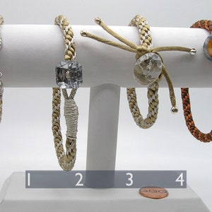 May include: Four braided bracelets with different closures and embellishments. The bracelets are displayed on a white stand. The bracelets are made of brown and beige cord. The first bracelet has a silver clasp. The second bracelet has a clear crystal square. The third bracelet has a clear crystal round bead. The fourth bracelet has a brown and orange button.