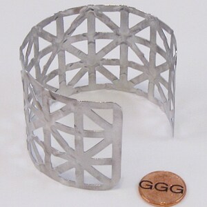 May include: A silver metal cuff bracelet with a geometric pattern. The bracelet is open on one side and has a textured surface. A penny is included for size reference.