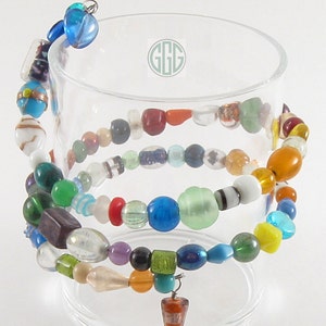 May include: Multicolored glass bead bracelet with a variety of shapes and sizes. The bracelet is wrapped around a clear glass cylinder. The bracelet has a small, colorful, cone-shaped charm.