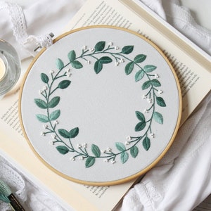 Op de afbeelding: Een witte borduurring met een ontwerp van een groene krans van bladeren. De krans is gemaakt van groene bladeren en witte bloemen.