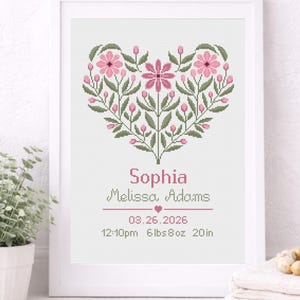 Puede incluir: Una obra de arte en punto de cruz enmarcada con un diseño floral en forma de corazón en rosa y verde. La obra incluye el nombre "Sophia", fecha, hora, peso (2,95 kg) y longitud (50,8 cm).