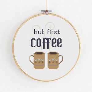 Op de afbeelding: Een witte borduurring met een kruissteekontwerp met de tekst "but first coffee" en twee bruine koffiemokken met stoom die eruit opstijgt.