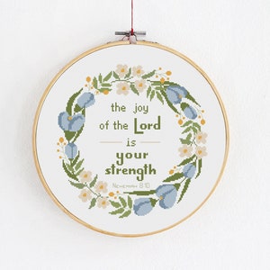 Könnte beinhalten: Ein Kreuzstich-Stickrahmen mit einem floralen Kranzdesign. Der Kranz besteht aus blauen, grünen und weißen Blumen. Der Text in der Mitte lautet "Die Freude des Herrn ist deine Stärke, Nehemia 8:10".
