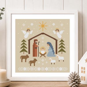 Puede incluir: Una pieza de arte en punto de cruz con marco cuadrado que muestra una escena de natividad pixelada sobre un fondo beige. El diseño incluye un pesebre central, figuras, dos ángeles, dos pinos y animales bajo una gran estrella amarilla.