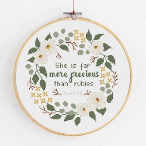 Może przedstawiać: Obręcz do haftu krzyżykowego z motywem kwiatowego wieńca. Wewnątrz napis: "Ona jest cenniejsza niż rubiny" z Przypowieści 31:10. Wieniec zawiera białe kwiaty, zielone liście i żółte akcenty.