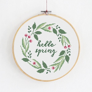 Puede incluir: Un bastidor de bordado de punto de cruz con un diseño de corona de primavera. La corona está compuesta por hojas verdes y bayas rosas, que rodean las palabras "hello spring" en una fuente de escritura. El bordado está en un bastidor de madera, sobre fondo blanco.