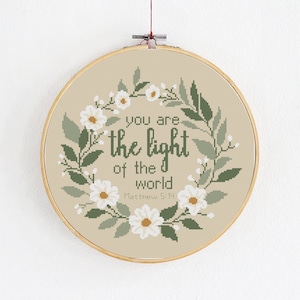 Può includere: Un cerchio da ricamo beige con un designo a corona di fiori e il testo "Voi siete la luce del mondo, Matteo 5:14".