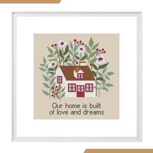 May include: Cross stitch pattern featuring a white house with a red door surrounded by green foliage and white flowers. The text "Our home is built of love and dreams" is below the house.