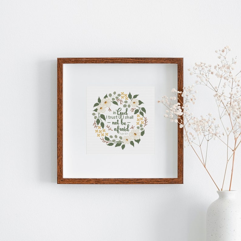 May include: A framed print with a brown wooden frame. The print features a floral wreath design with white flowers, green leaves, and the text "in God I trust I shall not be afraid". A white vase with flowers is in the background.