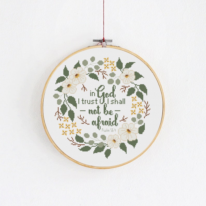 May include: A cross-stitch embroidery hoop featuring a floral wreath design. The text reads "in God I trust I shall - not be - afraid" from Psalm 56:4. The design includes white flowers, green leaves, and yellow accents.
