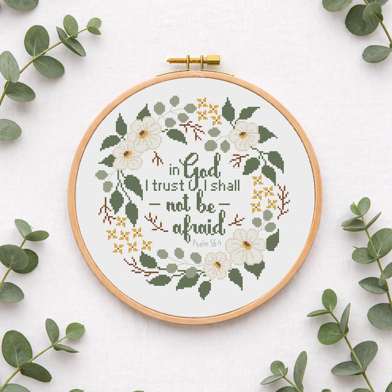 May include: A cross-stitch embroidery hoop featuring the text "in God I trust I shall not be afraid" from Psalm 56:4. The design includes white flowers, green leaves, and yellow accents, all set against a white background. The wooden hoop is visible.